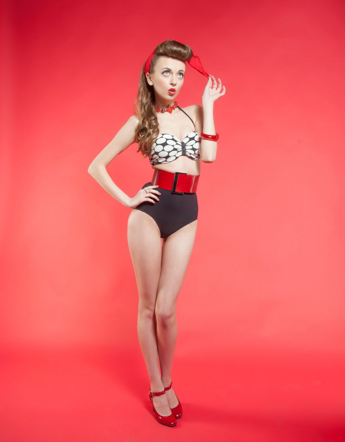 1940s pin up girl costume for Halloween A woman wearing a pin up costume for halloween, that has black bikini bottoms, a red belt, and a white top with black polka dots, here hair is pulled up, and she has a pin curl in the front, she is wearing red t strap shoes.