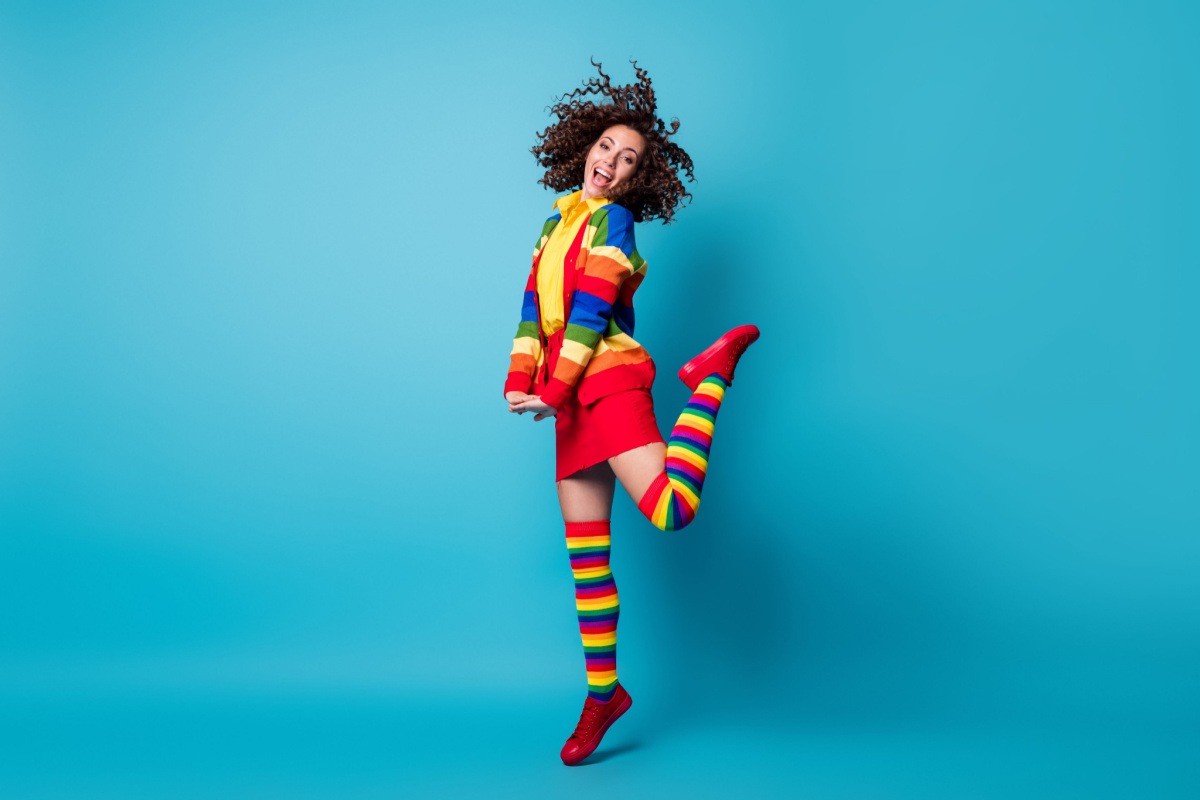 DIY rainbow costume A woman jumping with one leg bent at the knee behind her, wearing a DIY rainbow costume for adults which consists of knee high rainbow socks, a red skirt and a rainbow sweater over a yellow shirt.