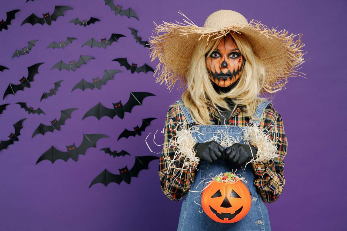 Scarecrow costume A diy scarecrow costume on a woman, consisting of denim overalls, and a plaid shirt, with straw coming out of her cuffs, and she is wearing a straw hat, with her face painted like a pumpkin, and she is holding a jack o lantern in front of a purple wall with bats.