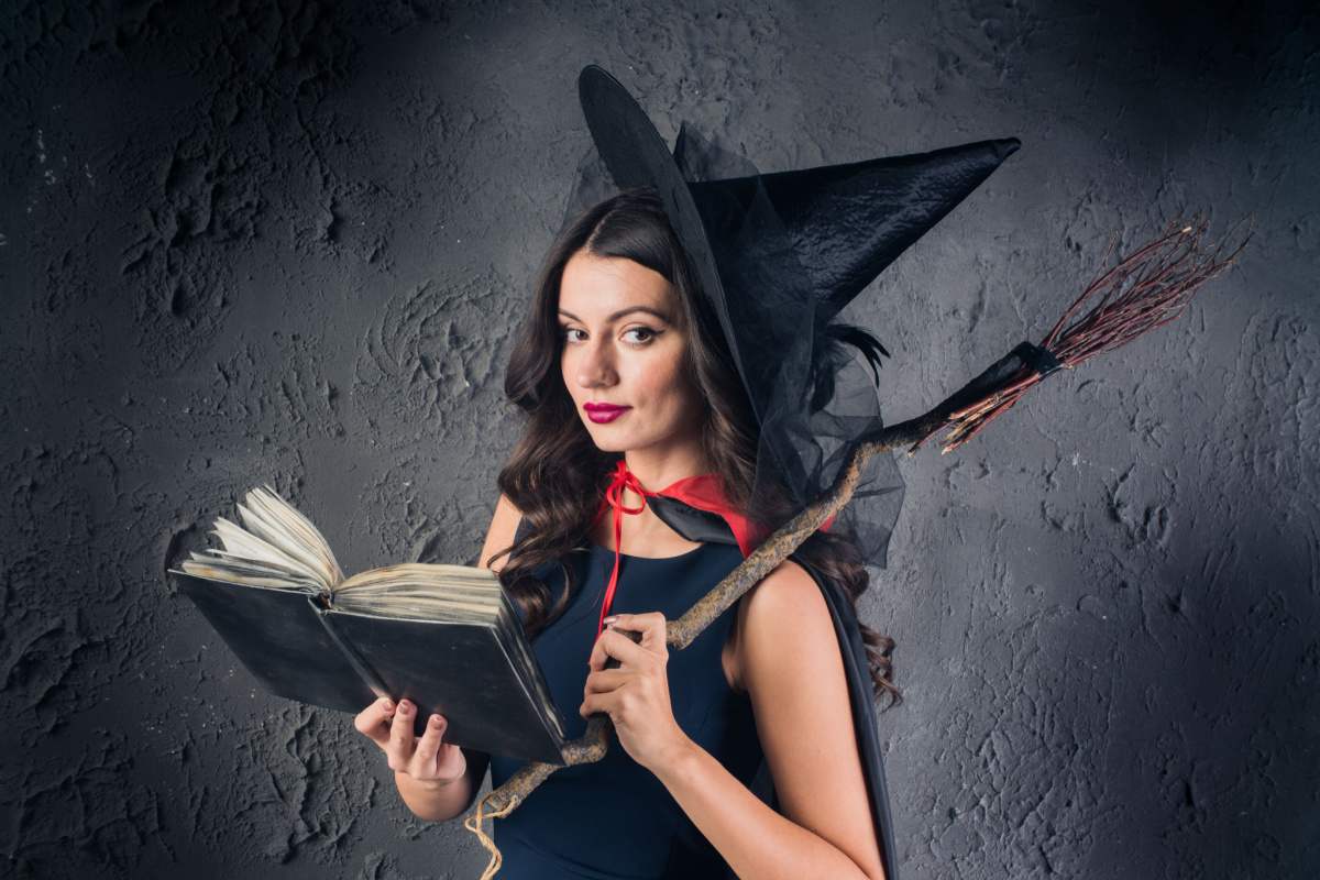 Witch costume A woman wearing a DIY witch costume for Halloween consisting of a black dress, a witches hat, a black cape with a red string around her shoulders, and she is holding a broom made of sticks, and a black spell book.