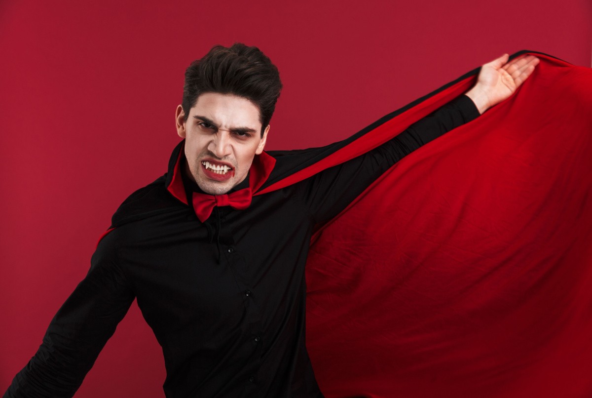 Vampire costume A man with dark hair wearing an easy vampire costume DIY idea for Halloween holding his cape open against a red background.