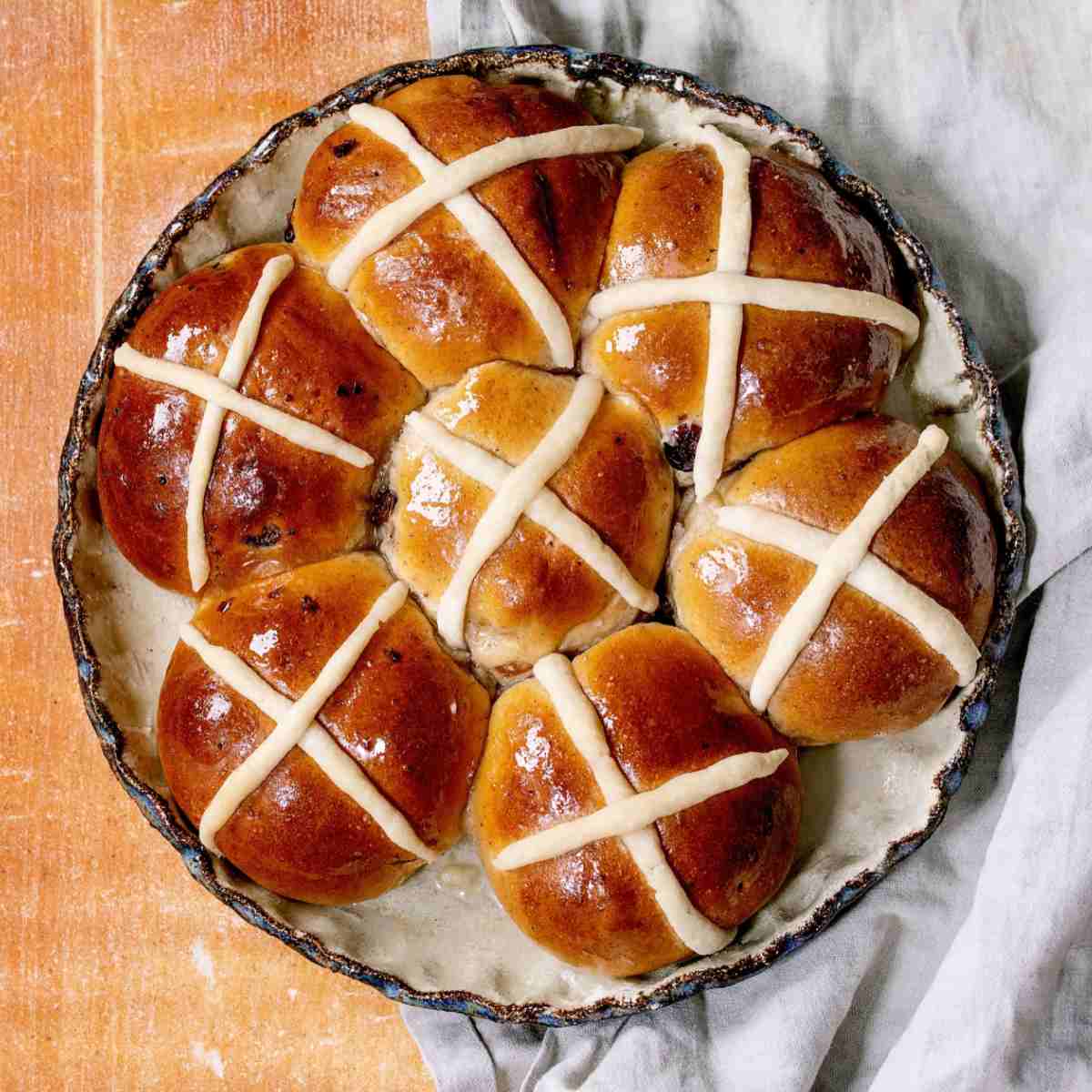 Hot cross buns in circular pie tin A pie tin of seven hot cross buns on a white cloth on top of a beige wooden backdrop.