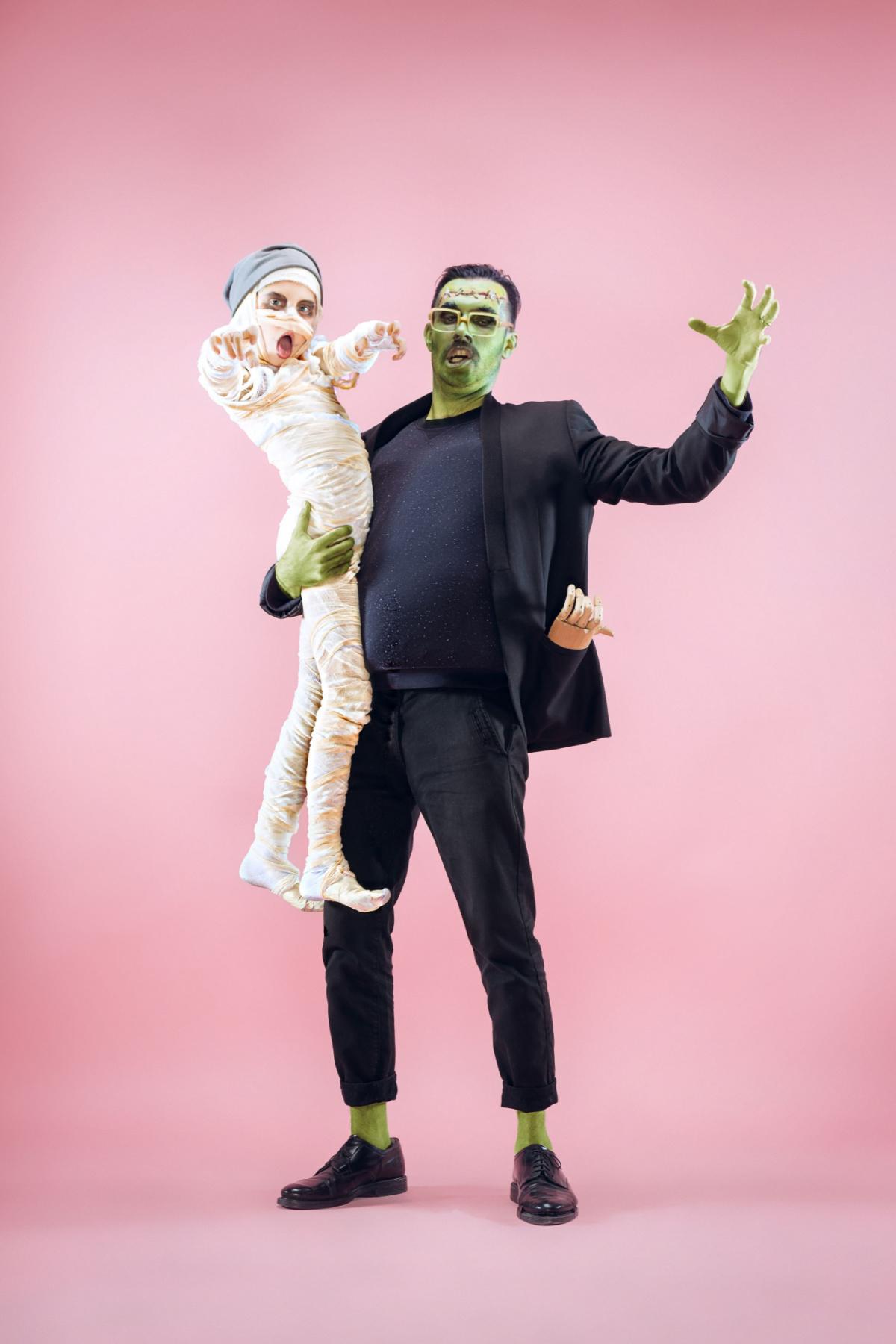 Guy in a Frankenstein costume holding a child in a mummy costume A man wearing a DIY Frankenstein costume holding a child wearing a diy mummy Halloween costume against a pink wall.