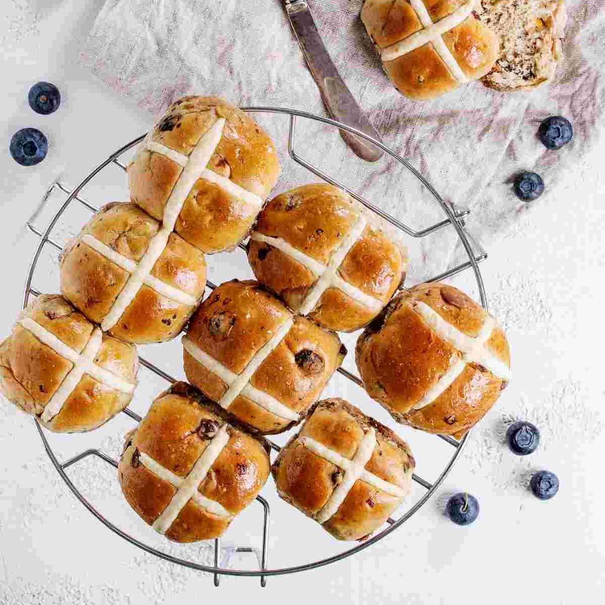 Hot cross buns on cooling rack A cooling rack of eight hot cross buns on National Hot Cross Bun Day with one hot cross bun on a cloth beside it, all of them are surrounded by blueberries.