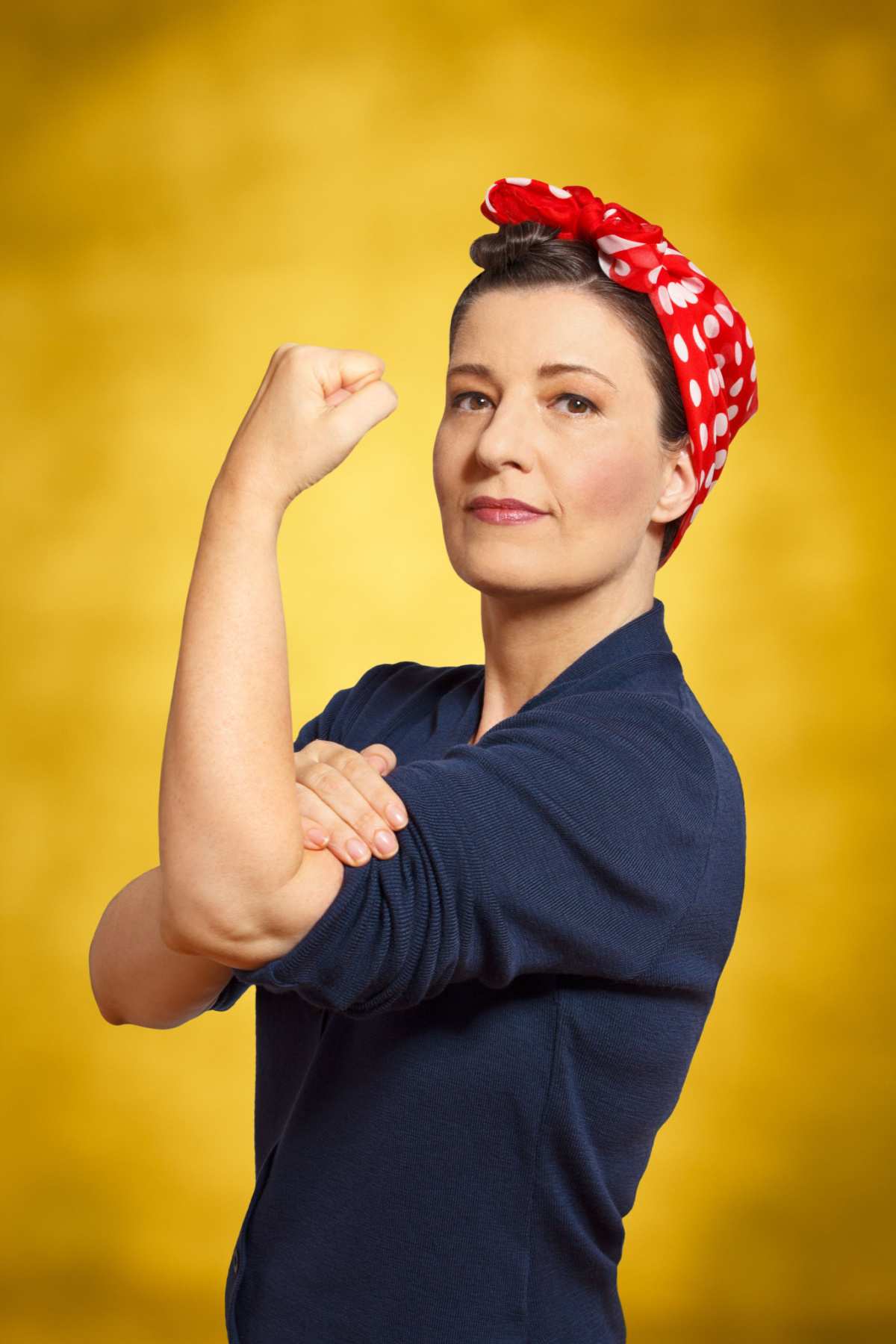 Rosie the Riveter costume DIY A Rosie the Riveter costume DIY idea on a woman curling her bicep in front of a yellow wall.