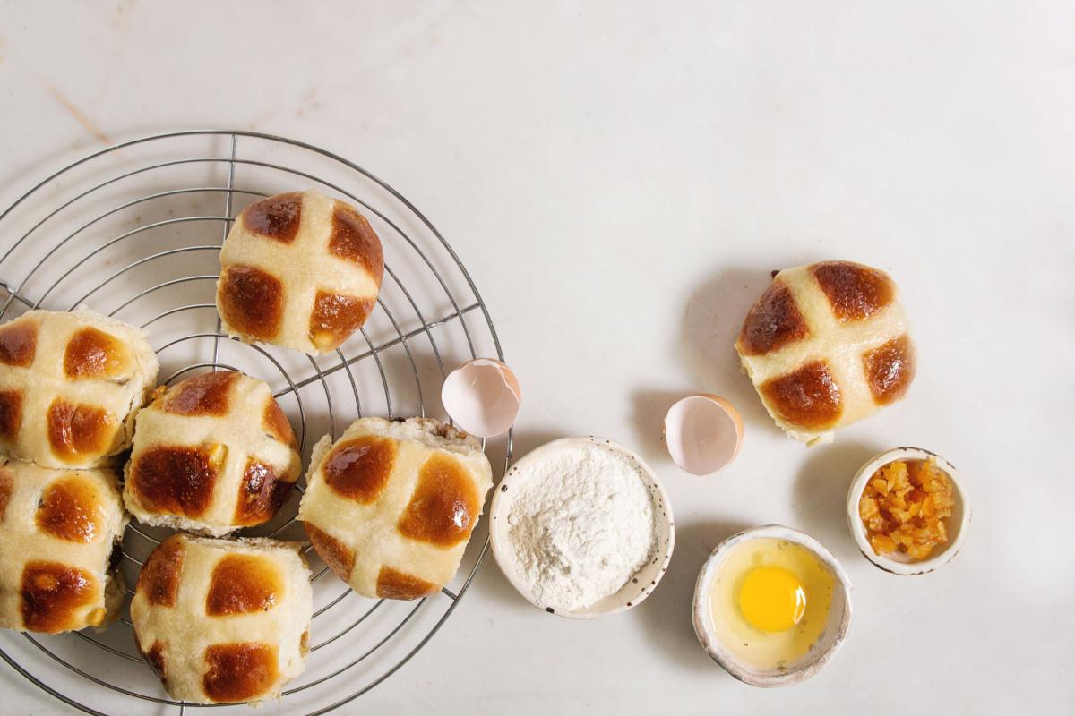 Hot cross buns and ingredients A cooling rack with homemade hot cross buns on it, with hot cross bun ingredients to the side, like eggs, flour, and golden raisins.