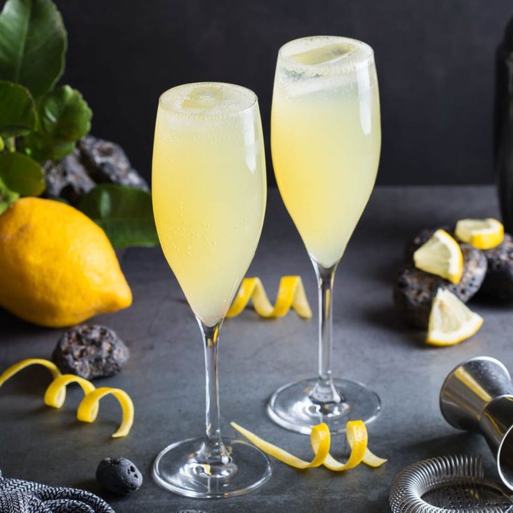 Two Champagne flutes filled with elderflower Prosecco cocktails, with lemon zest spirals, lemon slices, a jigger, and a lemon around them.