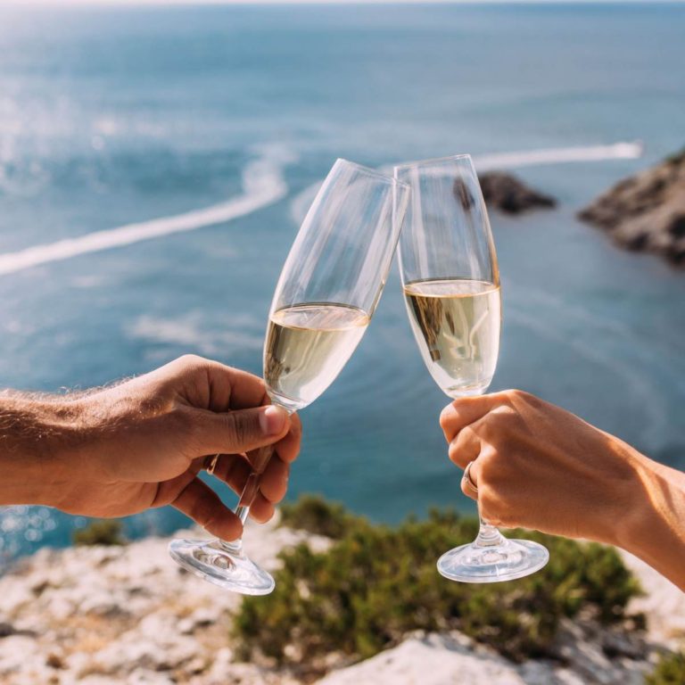 Two people clinking half filled champagne flutes together on a cliff overlooking a lake that has a speedboat driving by in the background.