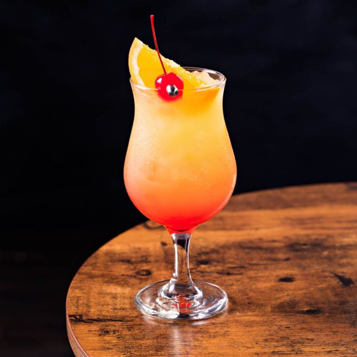 A hurricane cocktail garnished with a maraschino cherry and orange slice on a wooden table against a black background.