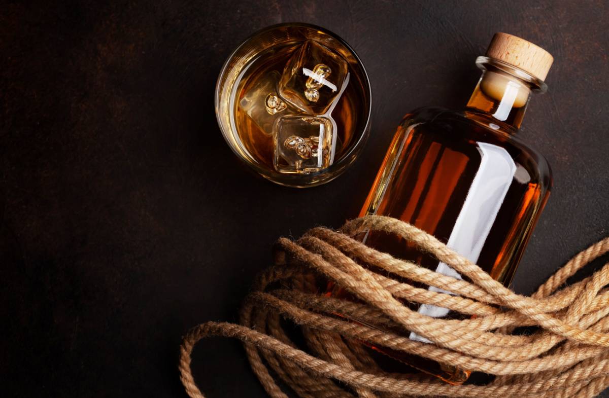 Navy rum bottle A glass bottle of naval rum wrapped in rope next to a rocks glass with ice and a pour of this naval strength rum in it.