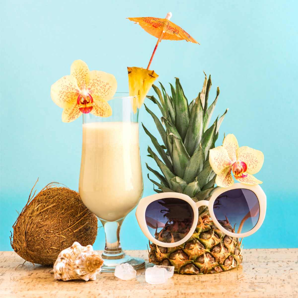 Pina colada and fruit A pina colada made with light rum garnished with an orchid flower, a pineapple slice and umbrella on a table next to a coconut and a pineapple half wearing sunglasses to look like a person.