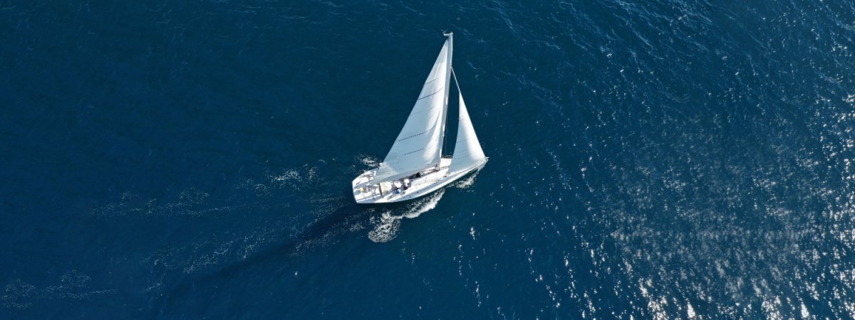 An overhead shot of a white sailboat with two sails moving eastwardly across the ocean.