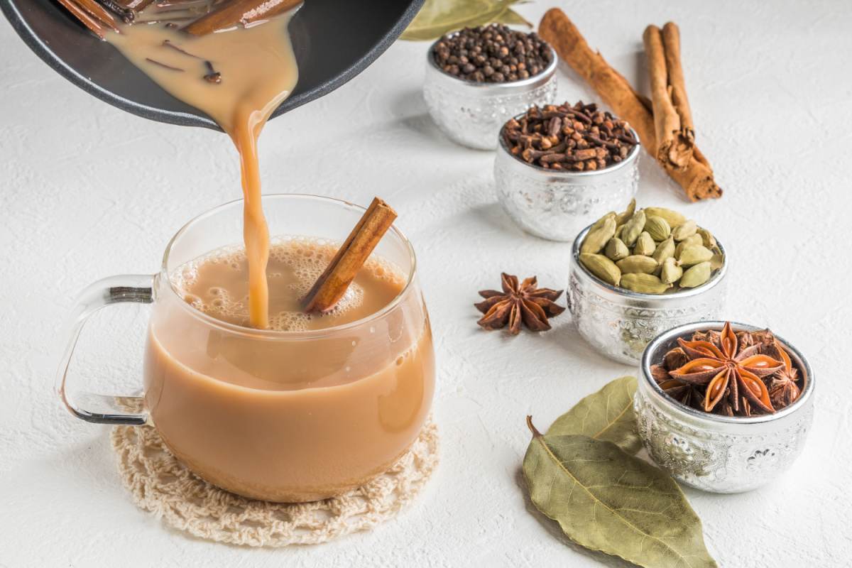 Pouring chai A saucepan filled with a homemade masala chai recipe being poured into a glass mug on a woven doily, with various chai spices in the background like star anise, green cardamom pods, black pepper, cinnamon sticks and cloves.