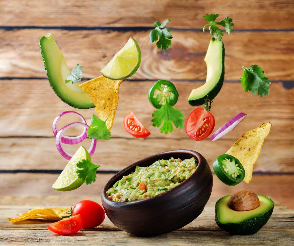 Flying guacamole ingredients A small bowl of homemade guacamole against a wooden background with guacamole ingredients floating above it, and a halved avocado beside it.