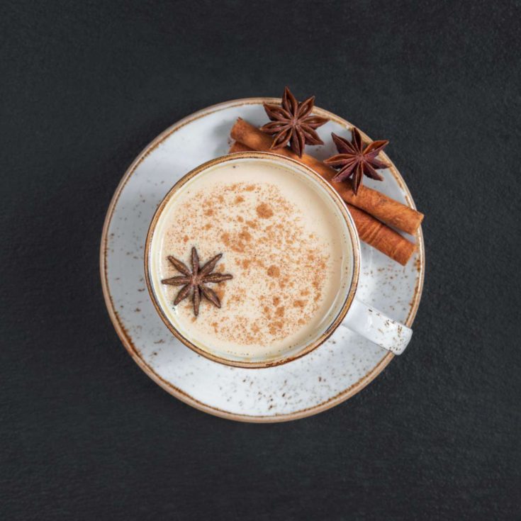 A white teacup and saucer filled with a homemade chai recipe garnished with one star anise in the cup, and two on the saucer along with two cinnamon sticks, all against a grey background.