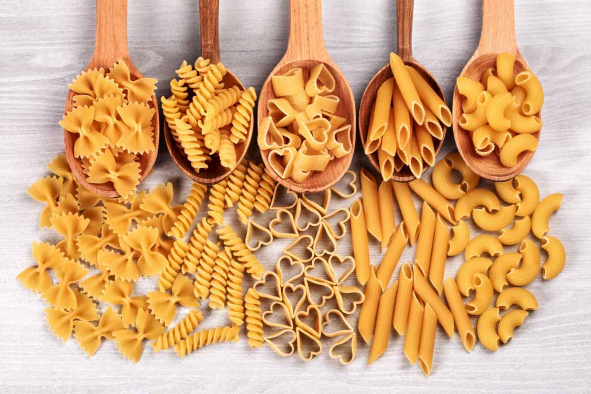 Short pasta types Five wooden spoons lined up in a row, with a different medium sized pasta types on each one, including penne, fusilli, macaroni, farfalle and heart shaped pastas.