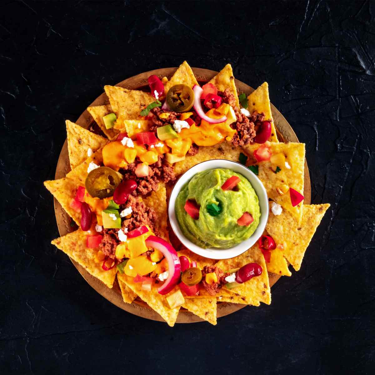 A circular plate filled with a homemade nachos recipe that has tortilla chips with ground beef, tomatoes, onions, jalapenos, cheese, kidney beans, avocado and corn on top, with a small ramekin of guacamole on top garnished with cilantro and more diced tomatoes. 