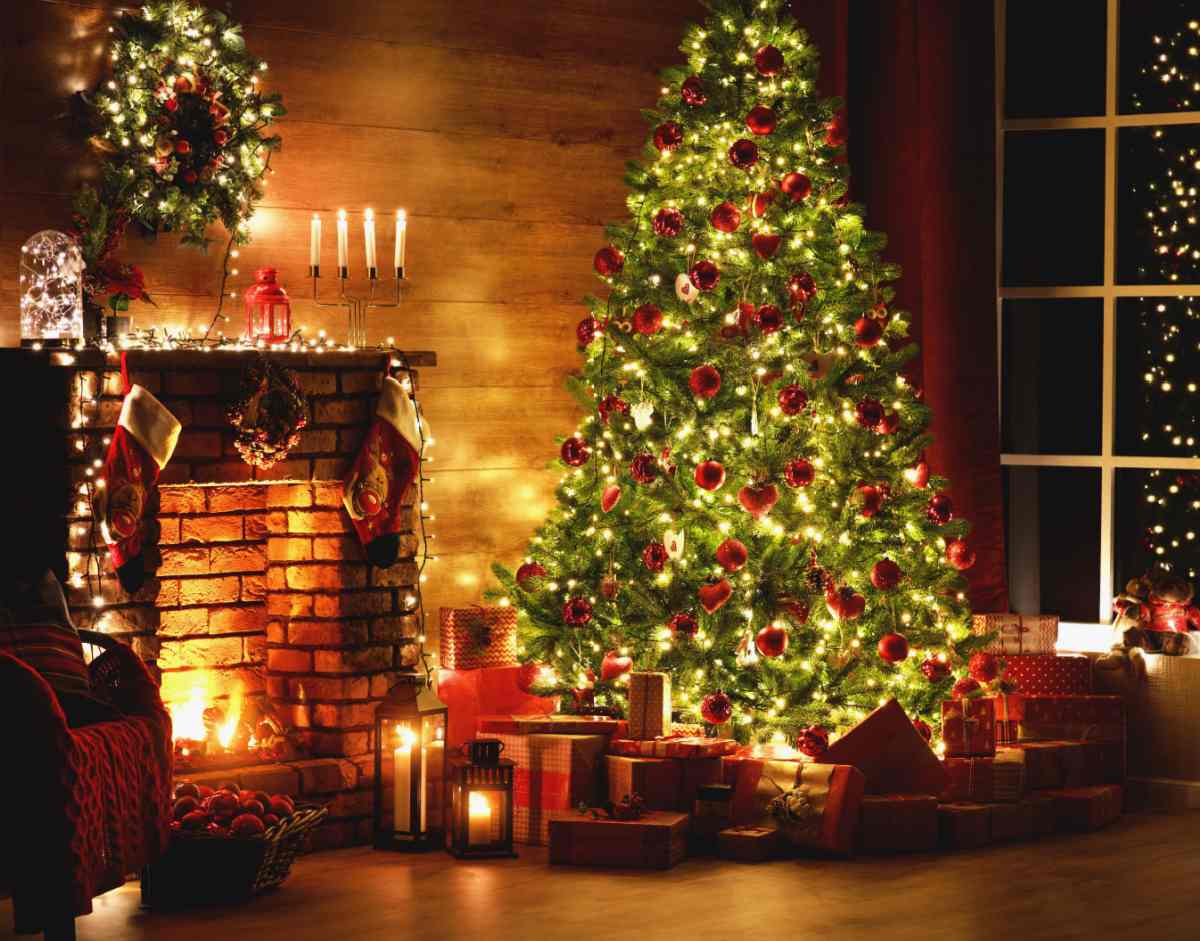 Christmas tree and fireplace A lit Christmas tree with red ornaments on it, next to a mantle with stockings hung on it, taper candles lit above it, and a fire burning inside the fire place.
