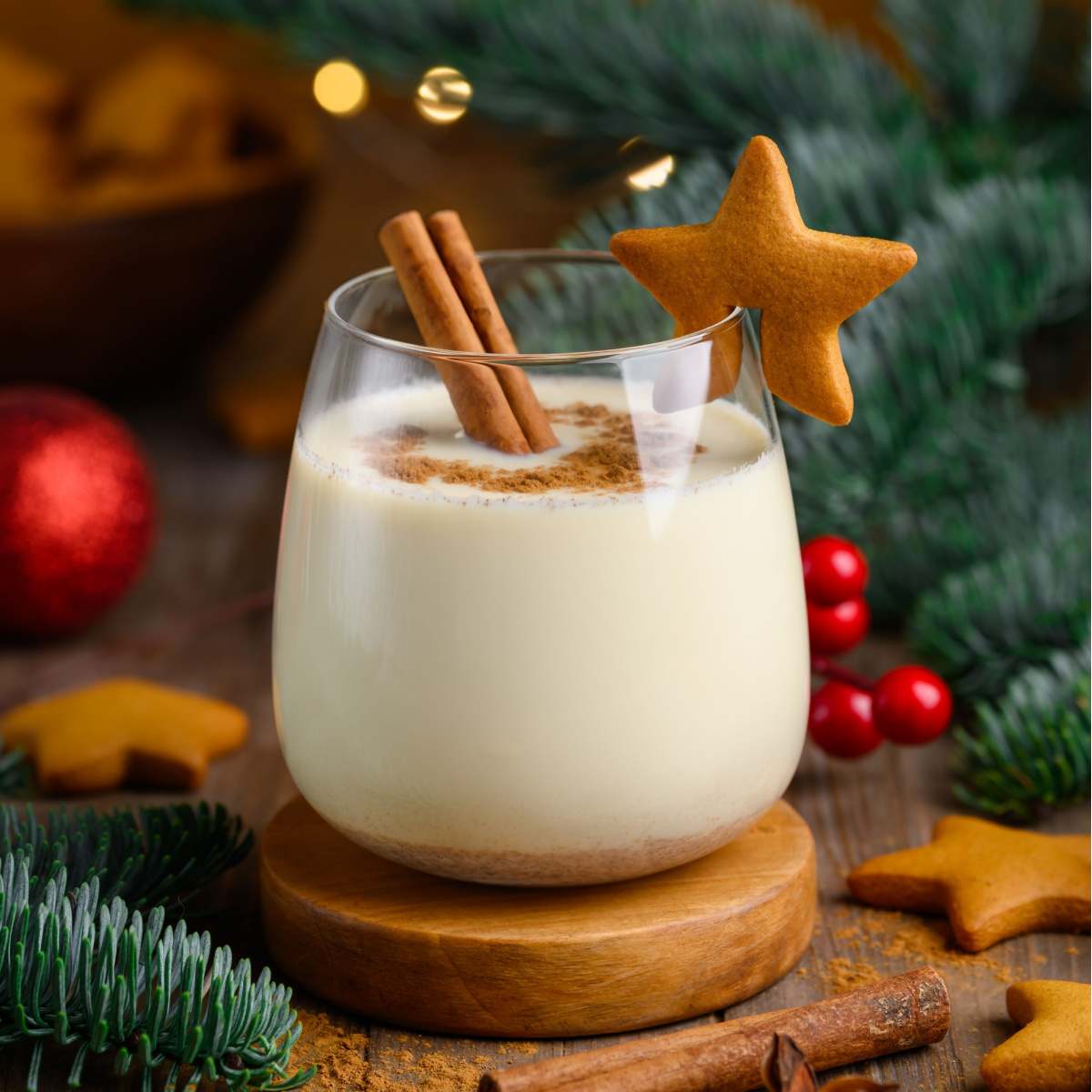 Glass of homemade eggnog A glass filled with a generous serving of homemade non alcoholic egg nog on a wooden coaster, garnished with a cinnamon stick and ginger bread cookie on the glass, surrounded by pine boughs, red Christmas ornaments, more gingerbread cookies, cinnamon sticks and star anise with a bokeh of lights in the background of the image.