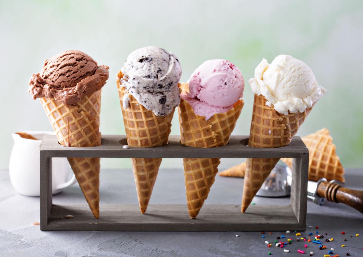 Four ice cream cones with different flavors of ice cream (from left to right: chocolate, cookies and cream, strawberry and vanilla) lined up in an ice cream cone holder for Ice Cream For Breakfast Day, with an ice cream scoop, extra waffle cone and small creamer in the background.