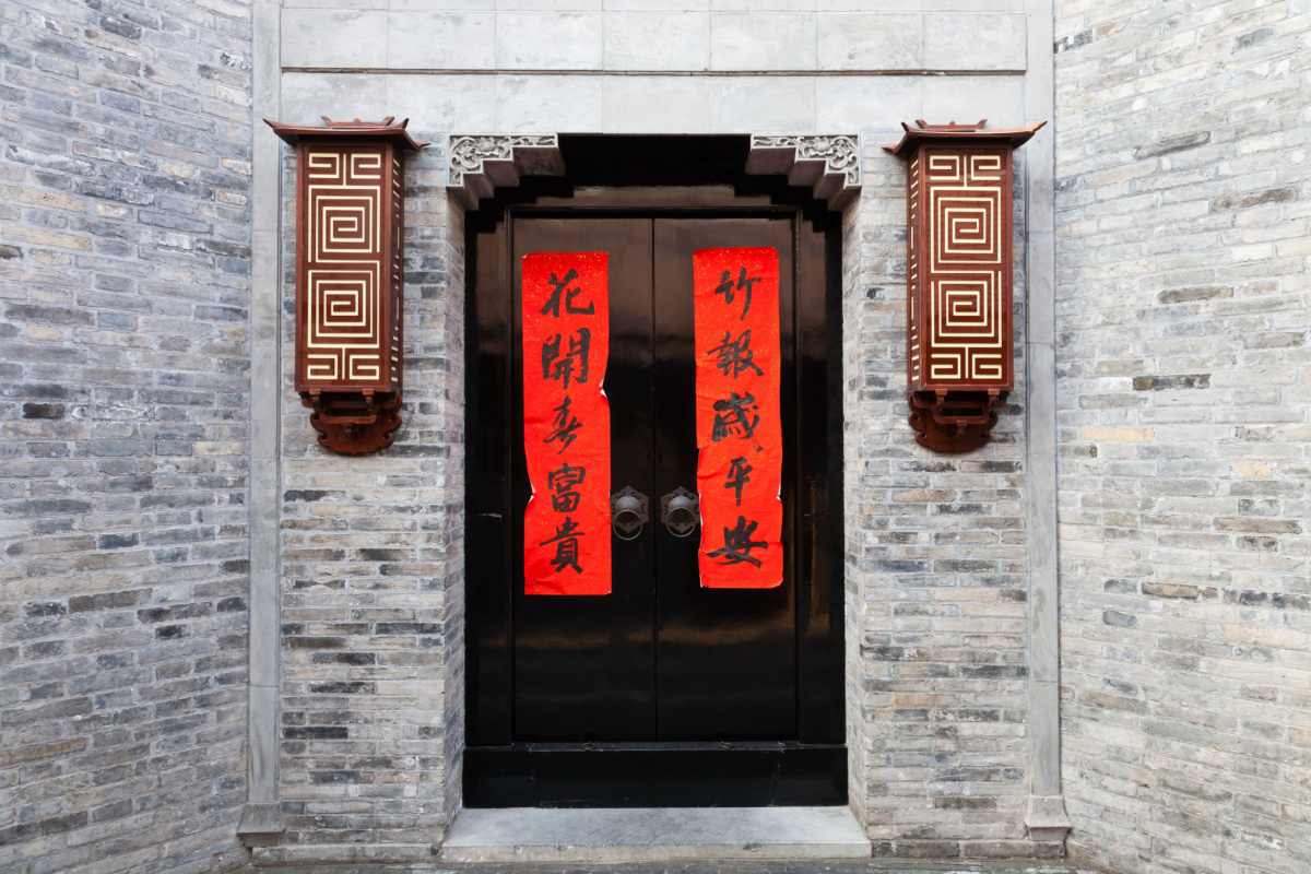 Chinese New Year couplets A grey brick wall with dark brown doors and two red Chinese New Year couplets taped to the door, with a light fixture on either side of the door.