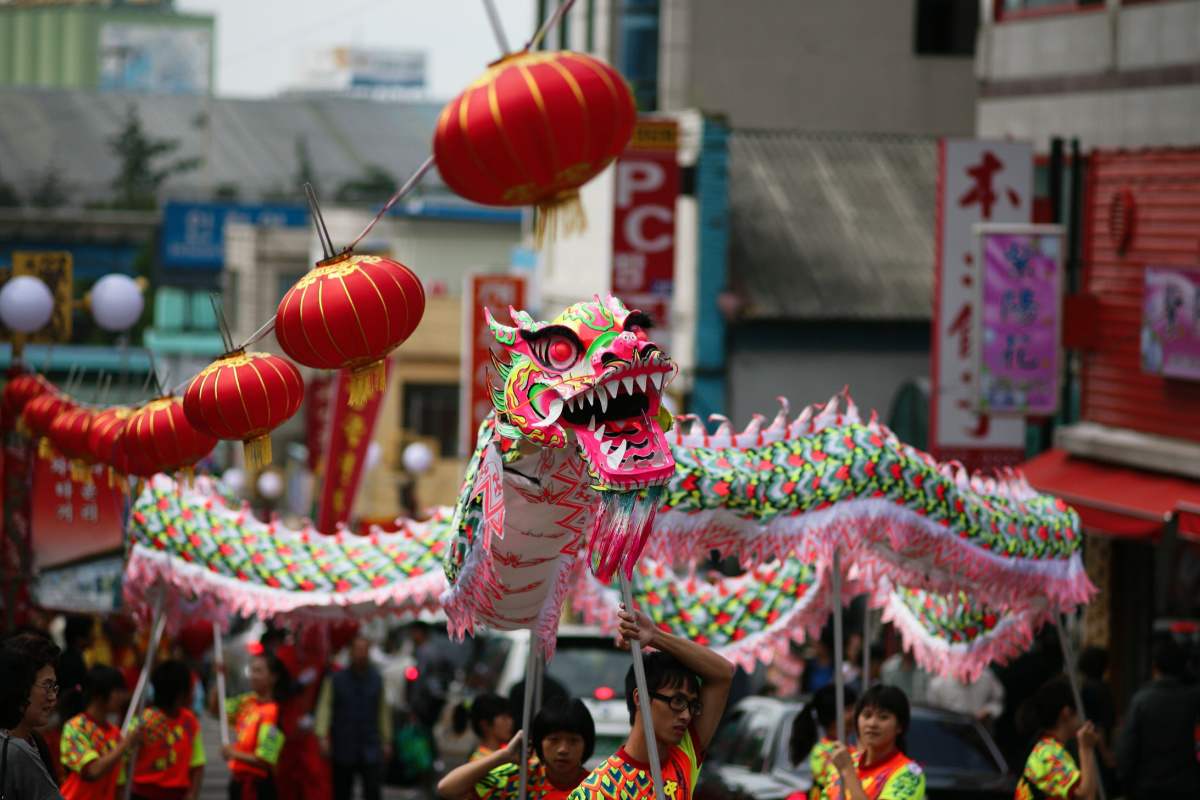 Chinese New Year dragon dance A Chinese New Year dragon dance being performed in a parade, with a green and pink dragon puppet, and red lanterns hanging overhead.