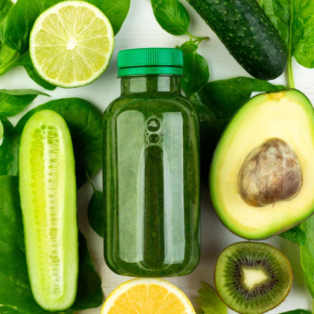 Bottle of green juice and veggies A close up shot of a bottle of cold pressed green juice containing an easy green juice recipe, resting atop various greens including spinach, limes, kiwi, avocados and cucumbers.