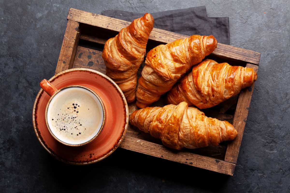 Box of croissants A cup of coffee in an orange cup on an orange saucer on top of an open wooden box with four freshly baked croissants in it to celebrate National Croissant Day.