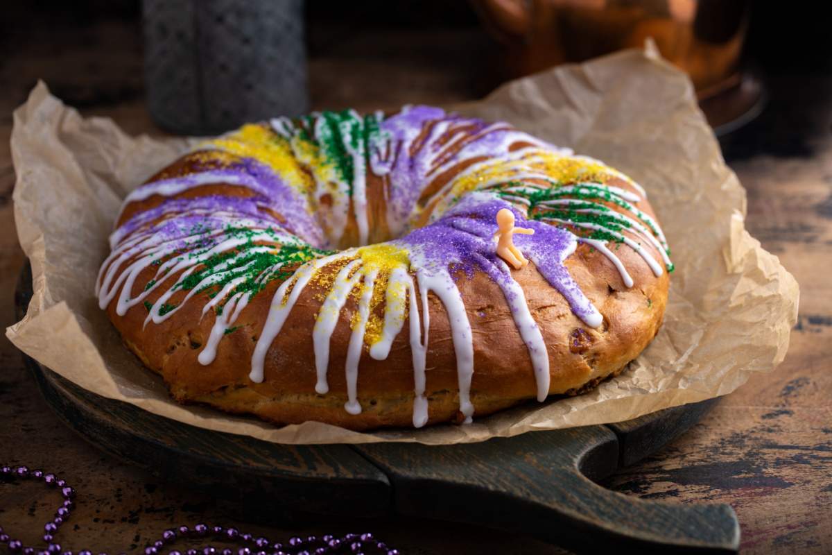 Mardi Gras king cake on parchment paper A Mardi Gras king cake with icing falling down the sides, and sprinkles in the colors of Mardi Gras on top of it, placed on top of a piece of parchment paper on top of a cutting board with a plastic king cake baby sitting on top of it.