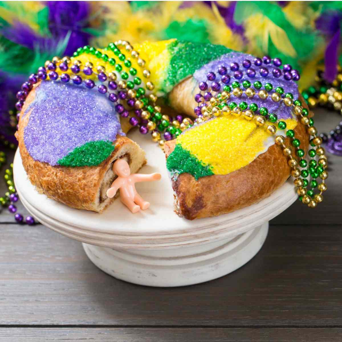 Mardi Gras king cake on a stand A Mardi Gras king cake on a cake stand with Mardi Gras beads draped on top of it, and a plastic king cake baby sitting in the space where a piece of cake was removed, with purple, green and gold yellow in the background.