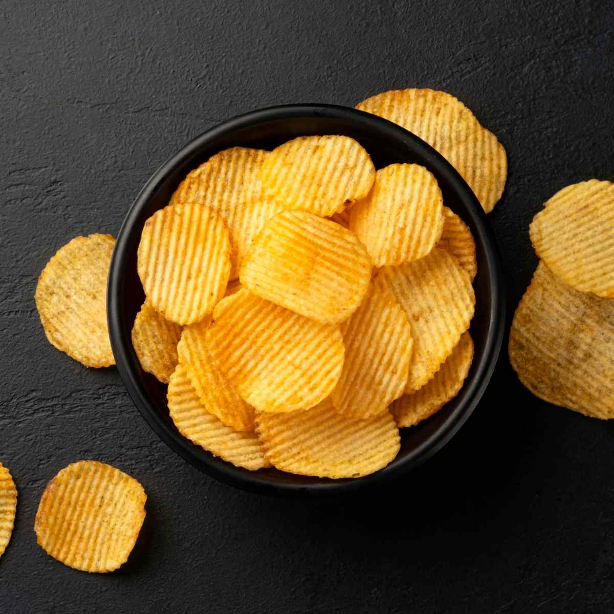 Bowl of potato chips A black bowl filled with ruffled potato chips on a dark brown background, with some of the potato chips spilling over the side and on the background tool.