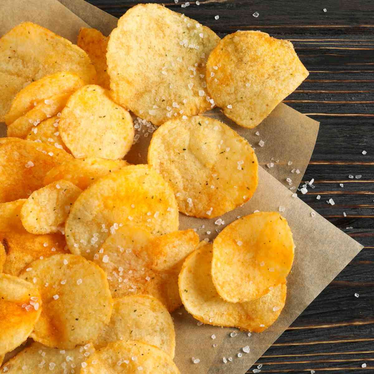Homemade potato chips on parchment paper A pile of homemade potato chips with sea salt piled on a piece of parchment paper on a wooden background.