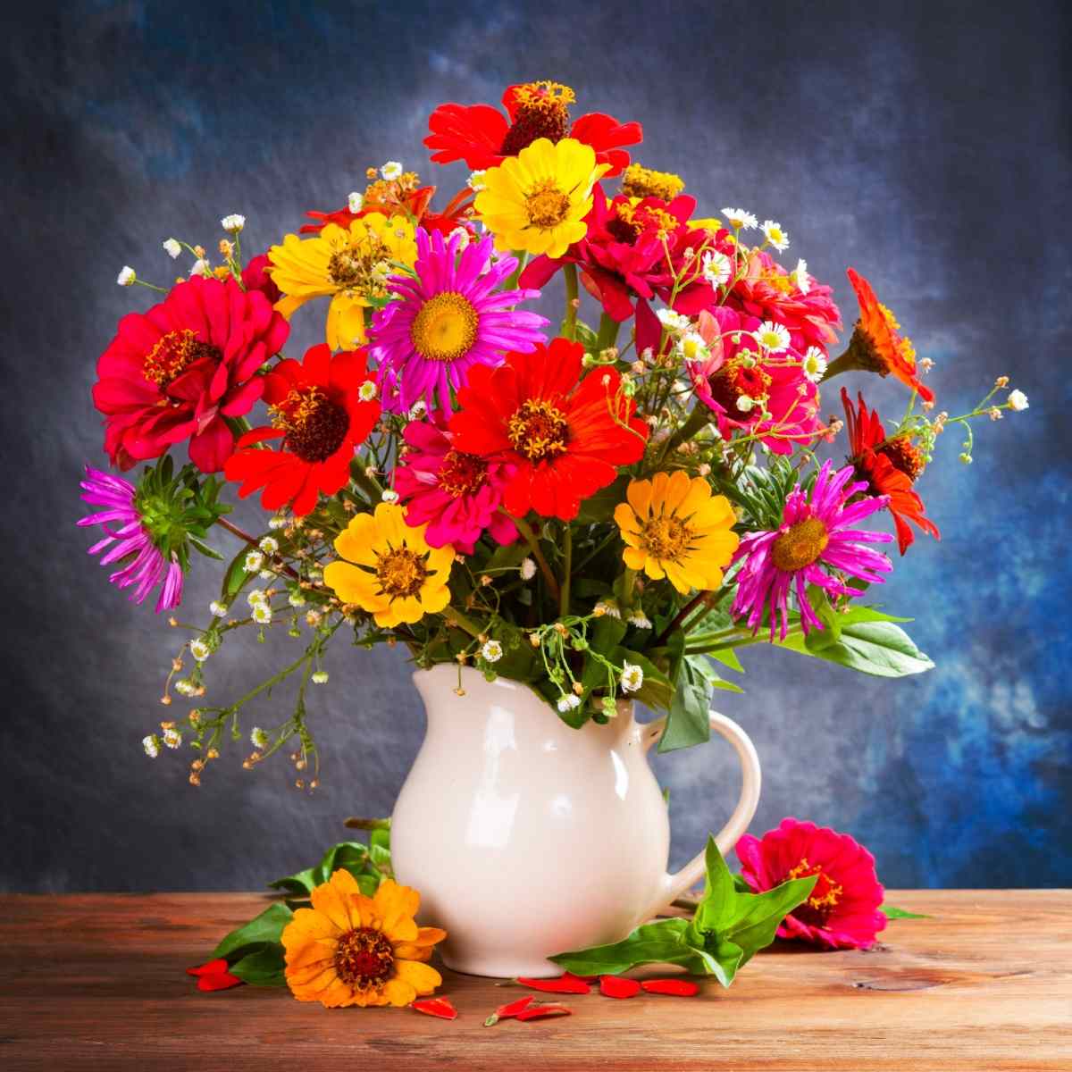 Flowers in a pitcher A white porcelain pitcher being used as a vase sitting on a wooden table against a blue background with various wildflowers in red, yellow, purple and white inside of it.