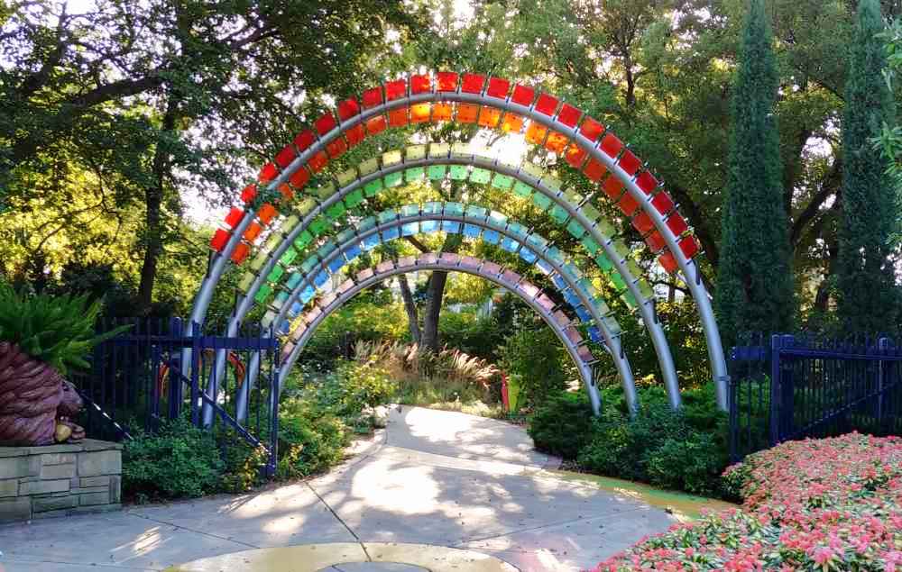 A photo of the rainbow arch that marks the entrance to the children's garden in Botanica, the Wichita Botanical garden, with light coming through the trees behind it, casting shadows onto the sidewalk, with a bush of pink flowers in the foreground.