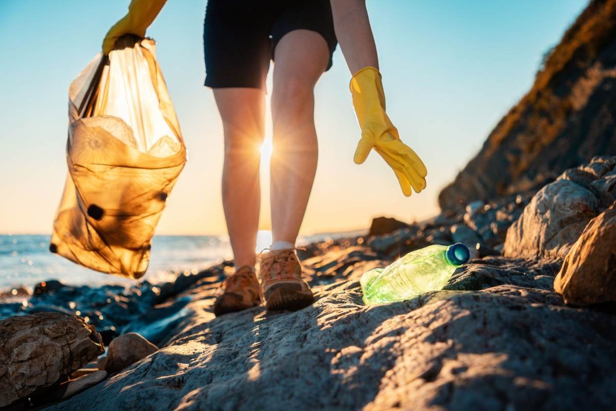 A woman picking up trash for Earth Day who is walking along a rocky coastline reaching for a green recyclable bottle, to put in the bag of bottles she's carrying with her.