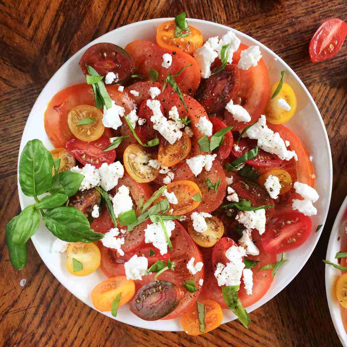 A caprese salad recipe featuring heirloom tomatoes, basil, mozzarella cheese, salt and pepper on a white plate sitting on a wooden coffee table.