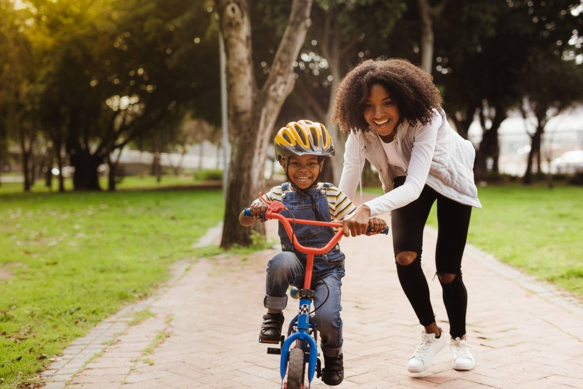 A mother helping her son who is learning to ride a bicycle down a path, by holding on to his shoulders gently to guide him as he begins to pedal away.