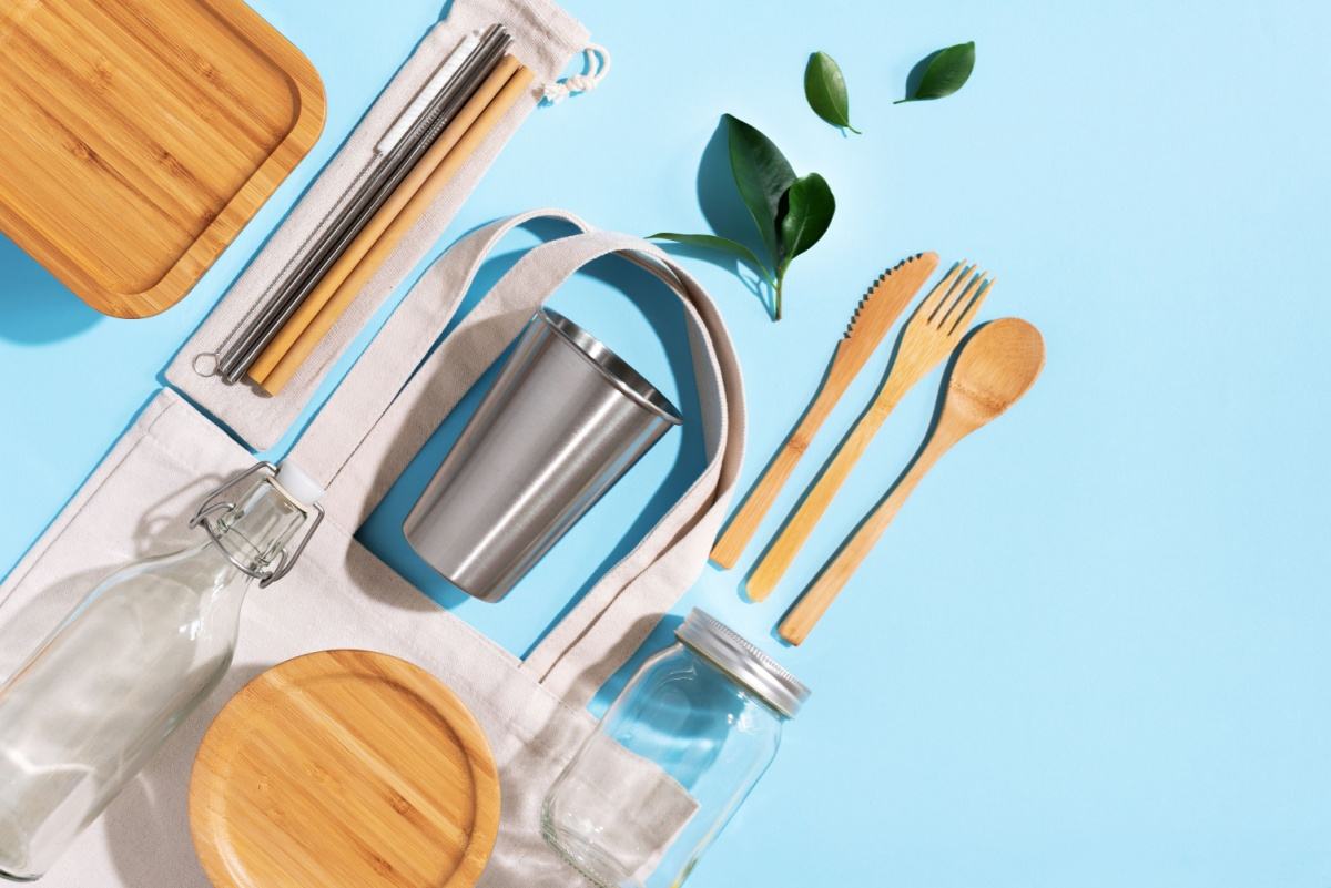 A flatlay image of reusable items including wooden cutlery, a metal cup, a cottom bag, bamboo plates, a glass jar, and a reusable metal straw, showing how to celebrate Earth Day by using reusable goods.