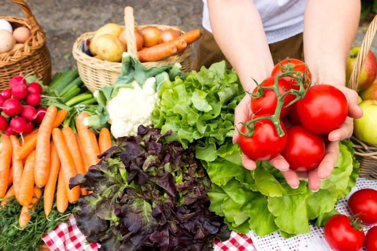 A woman's hands holding a bunch of tomatoes on the vine, with lettuce, carrots, potatoes and radishes, visible in the background on a table at the farmer's market.