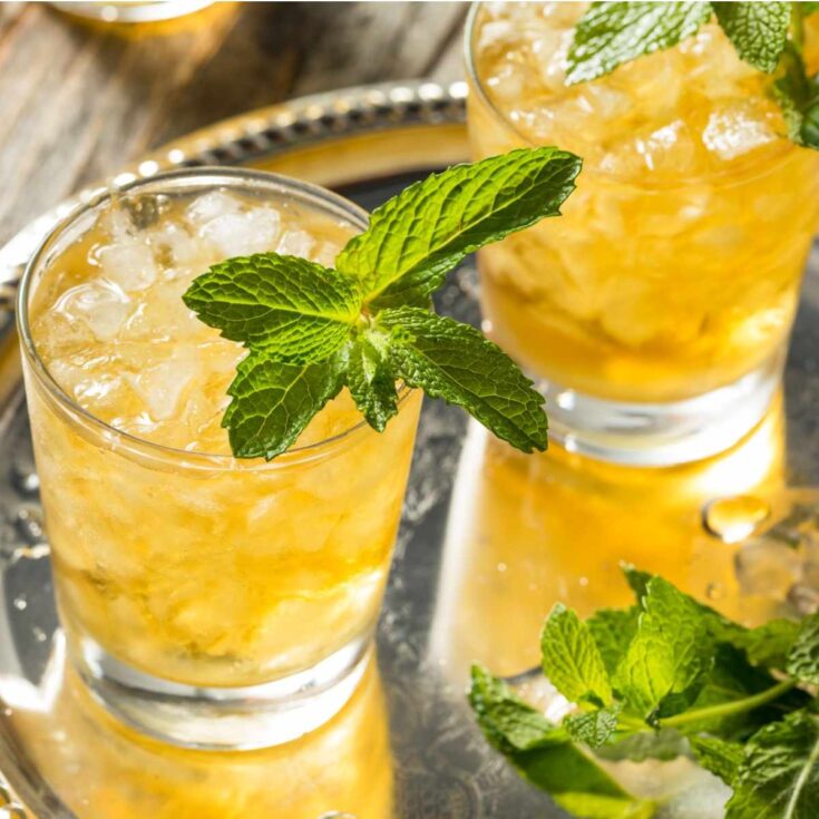 Two mint juleps in clear rocks glasses, garnished with mint on a silver serving tray with mint in the foreground to celebrate National Mint Julep Day.