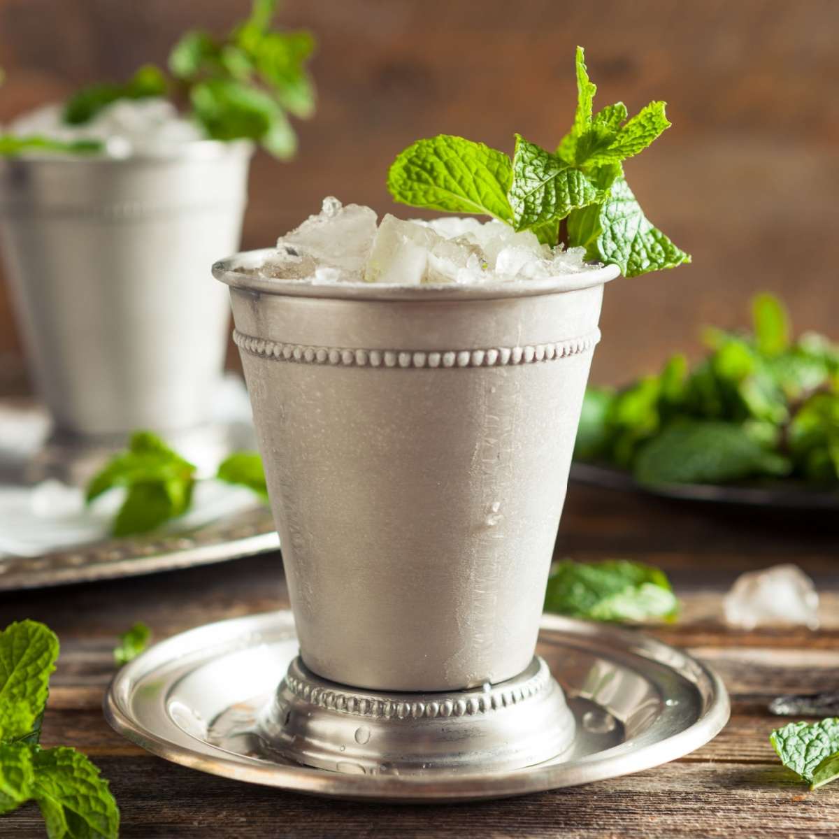 Mint julep in silver mug A mint julep garnished with mint on a silver saucer, in a traditional mint julep silver cup, with another mint julep and fresh mint in the background.