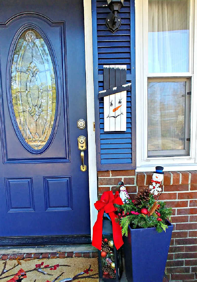 DIY snowman decor project on a blue shutter near a decorated front porch for the holidays.