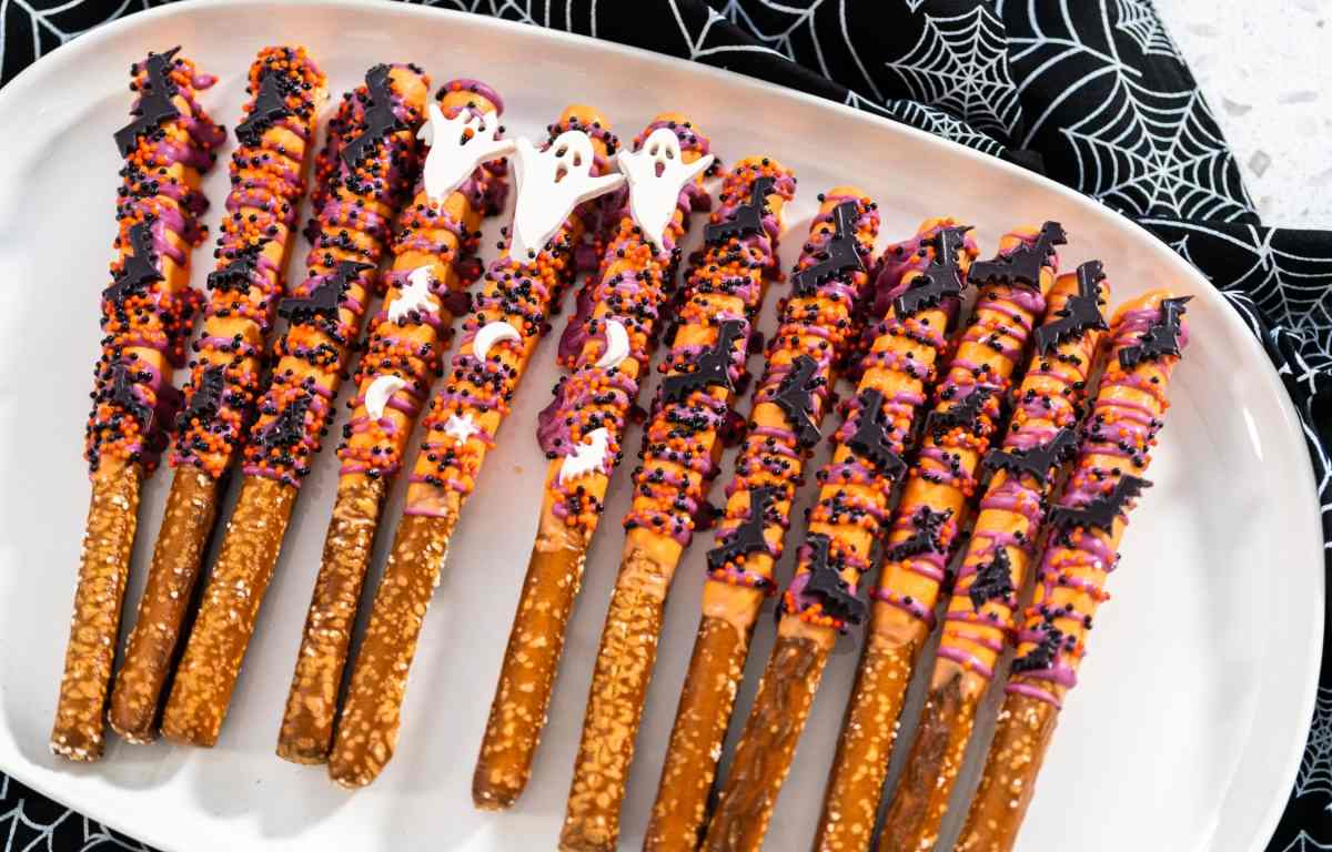 A white plate filled with Halloween pretzel rods decorated with melted chocolate, bat sprinkles, and ghost sprinkles.