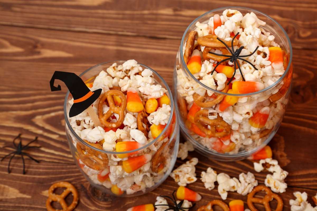 Two rocks glasses filled with a Halloween pretzel snacks mix with pretzel twists, candy corn and popcorn, each glass is garnished with either a plastic spider or a witches hat cocktail pick.