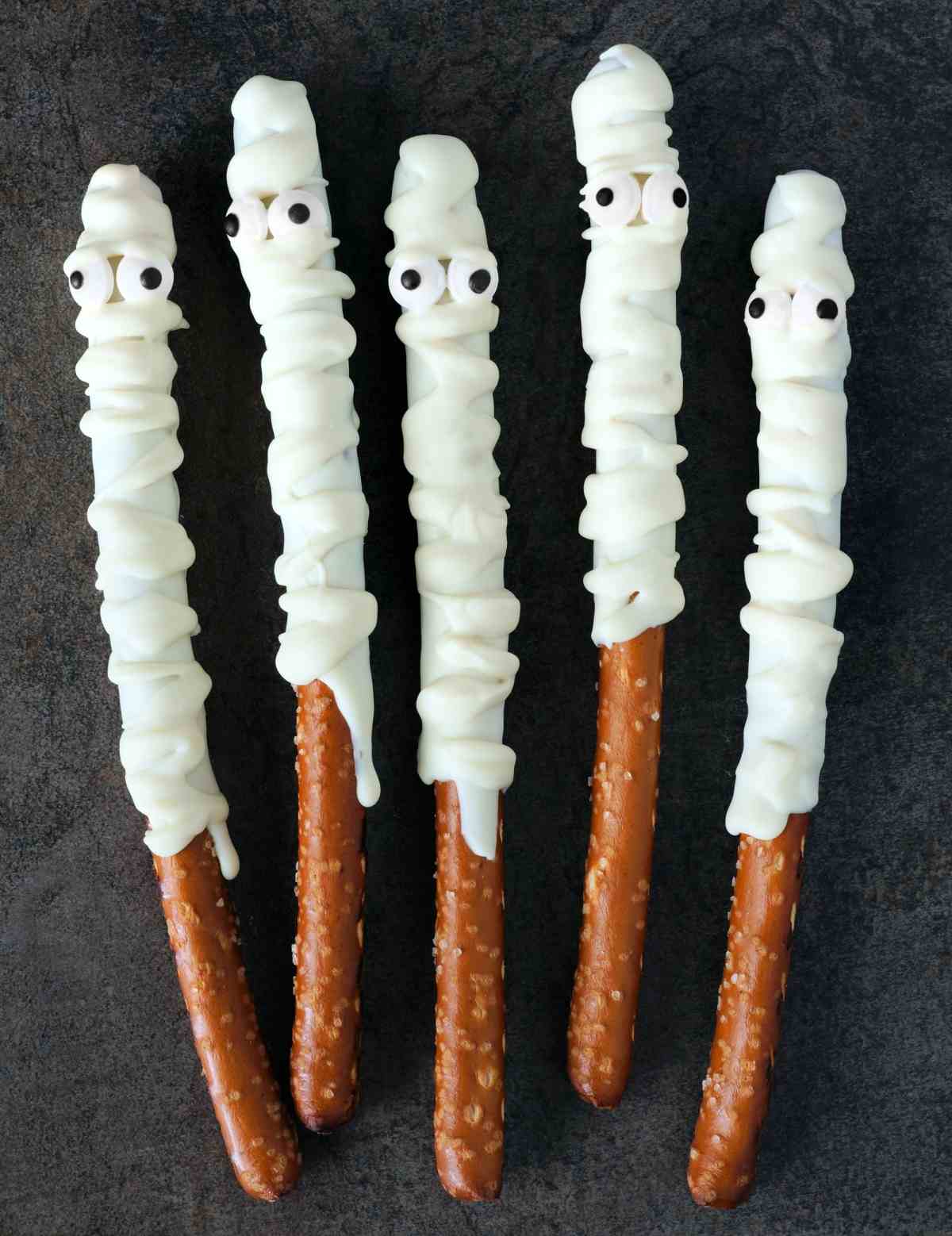 Five mummy pretzels with white chocolate drizzle and candy eyes decorating each of the mummy pretzel rods.