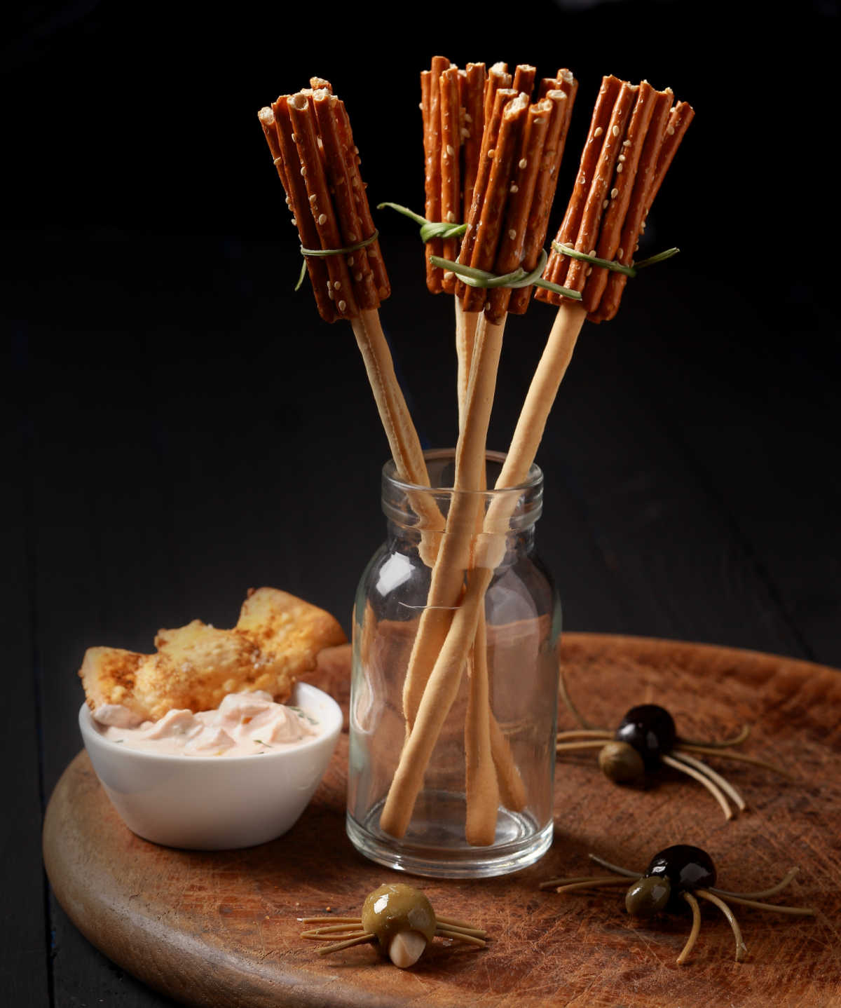A clear jar holding four pretzel broomsticks next to a spider made out of olives and a small bowl of crunchy bar snacks.