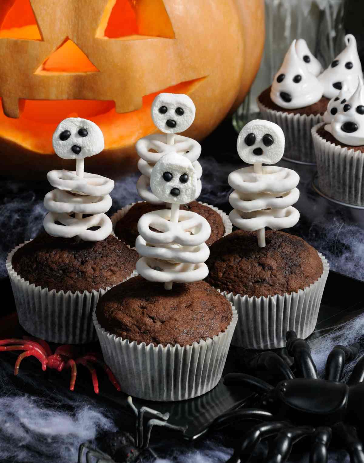Four pretzel skeleton cupcakes on a decorated Halloween party table, with yogurt pretzels and marshmallows making up the body of the skeleton pretzel cupcakes toppers.